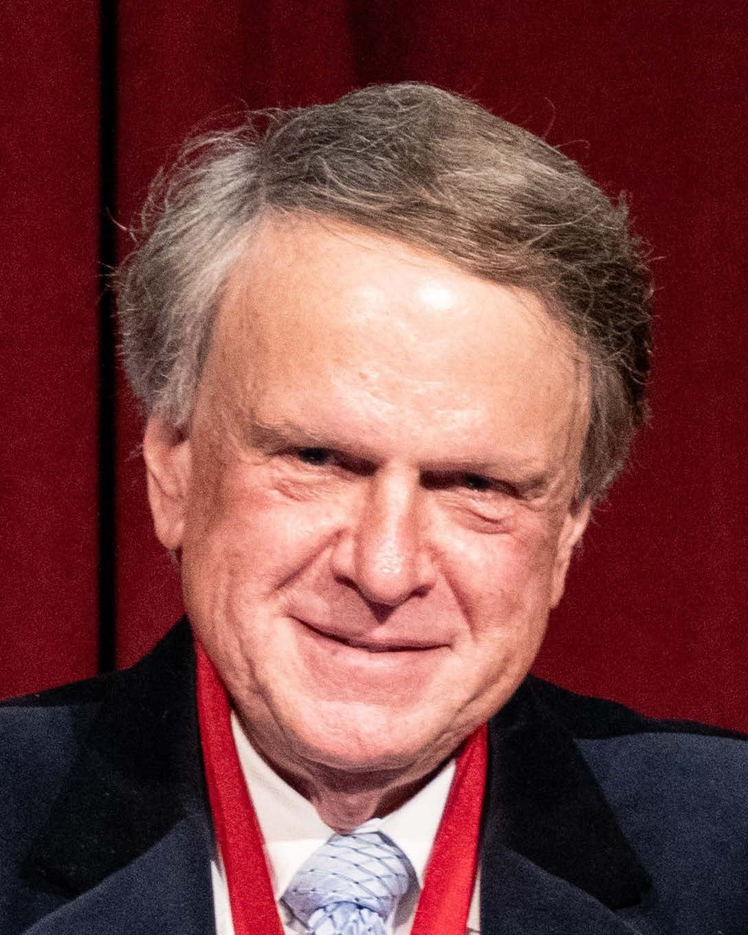Headshot of John Baras, a man smiling wearing a dark suit, white shirt, and blue tie