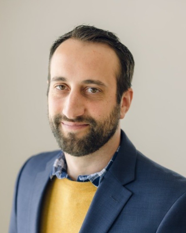 Headshot of Lee Greely, a man smiling wearing a blue jacket and yellow shirt