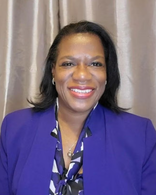 Headshot of Lora Brown, a woman smiling wearing a purple jacket and shirt