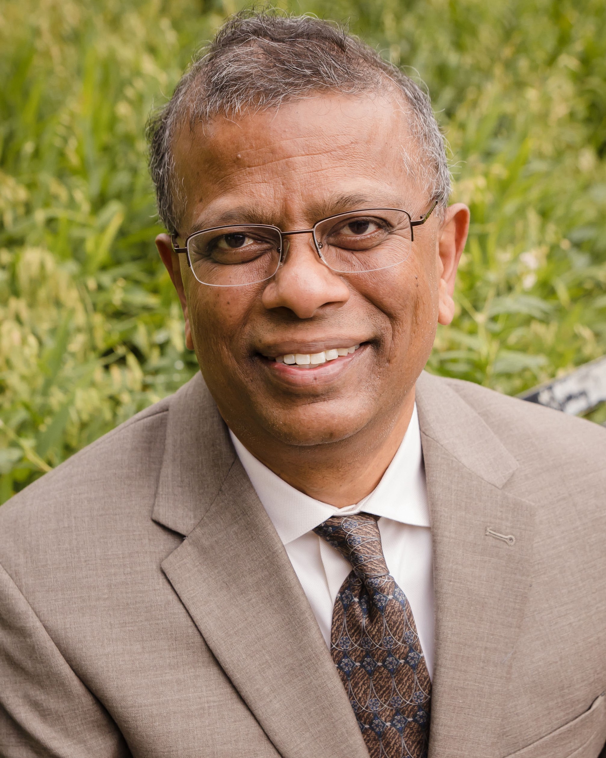 Headshot of Suresh Babu, a man smiling and wearing glasses, a taupe colored suit, and a brown tie
