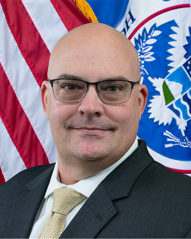 Headshot of Wayne Dumais Headshot of Wayne Dumais, smiling and wearing black frame glasses, a dark suit jacket, and a tan tie