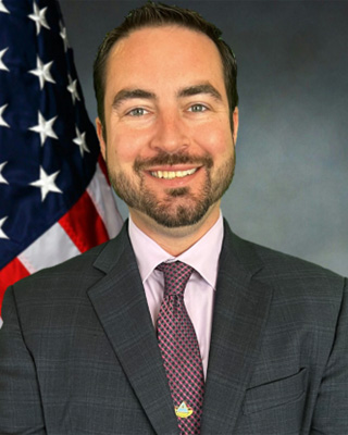 Headshot of Charles Rea, a man with dark hair and beard smiling and wearing a dark jacket, pink shirt, and purple tie