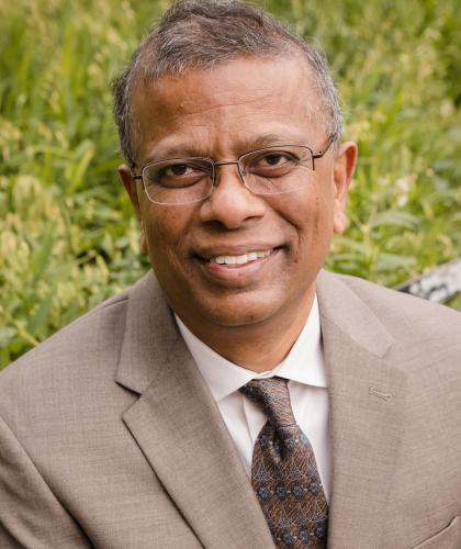 Headshot of Suresh Babu, a man smiling and wearing glasses, a taupe colored suit, and a brown tie